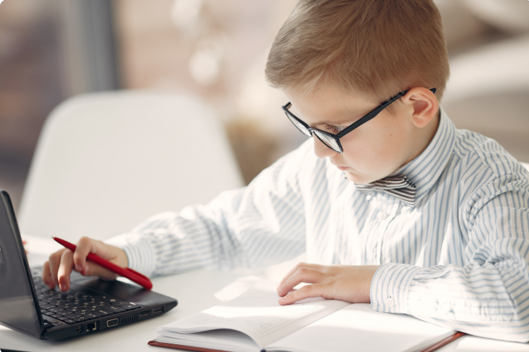 A young studious 2E boy at a computer, wearing a bow tie and glasses, with a notebook in front of him and a pencil in in his hand.