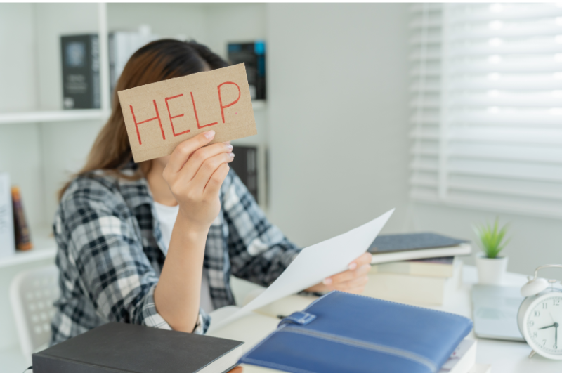 A stressed woman sitting at her desk, surrounded by papers and books, holding a sign over her face that says "Help," illustrating the struggles with overachievement.